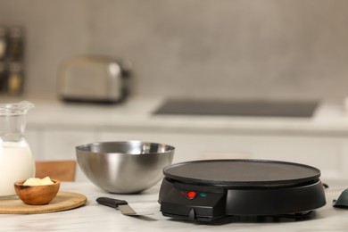 Photo of Electrical crepe maker and ingredients on white marble table in kitchen, space for text