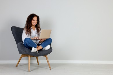 Young African-American woman working on laptop in armchair indoors. Space for text