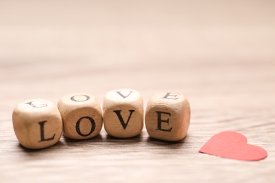 Mini cubes with letters forming word Love near red paper heart on wooden background, closeup