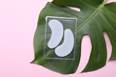 Package with under eye patches and green leaf on light pink background, flat lay. Cosmetic product