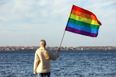 Woman holding bright LGBT flag near river, back view