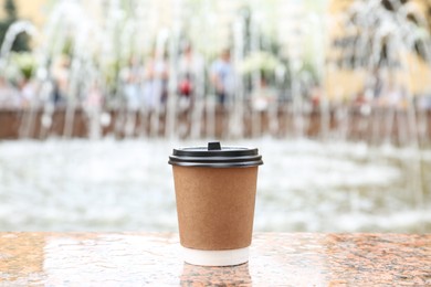 Photo of Cardboard cup with plastic lid on stone parapet near fountain outdoors. Coffee to go