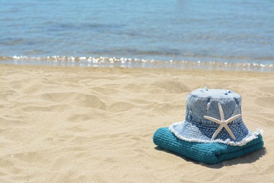 Stylish denim hat, towel and starfish on sand near sea, space for text. Beach accessories