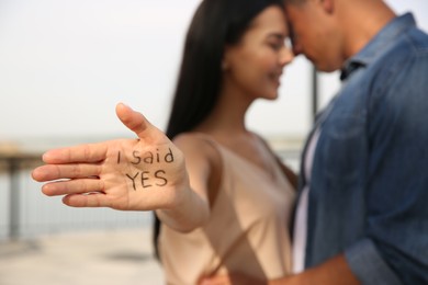 Photo of Woman with text I SAID YES written on palm and her boyfriend after engagement outdoors, focus on hand