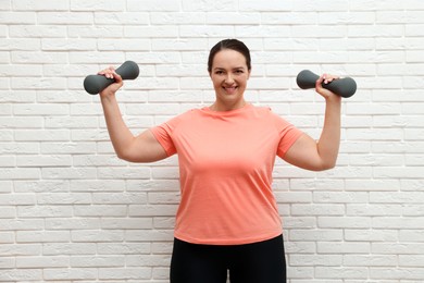 Happy overweight woman doing exercise with dumbbells near white brick wall