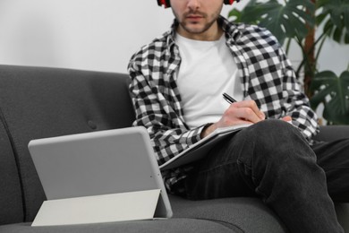 Photo of Young man with modern tablet and headphones studying on sofa at home, closeup. Distance learning