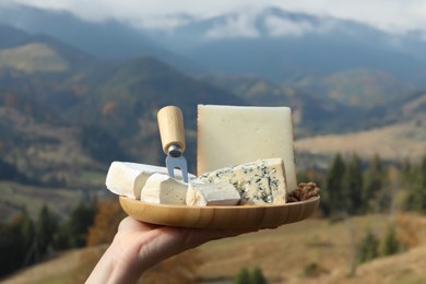 Woman holding wooden plate with different types of delicious cheeses against mountain landscape, closeup