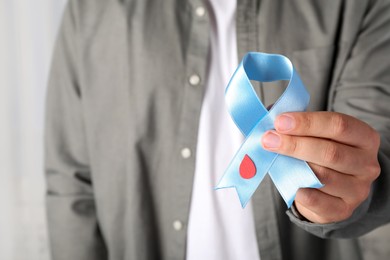 Man holding light blue ribbon with paper blood drop indoors, closeup. World Diabetes Day
