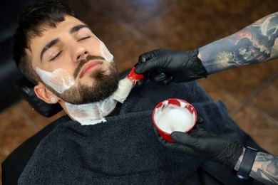 Photo of Professional hairdresser applying shaving foam onto client's beard in barbershop