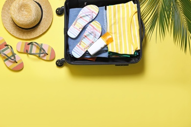 Open suitcase and beach accessories on yellow background, flat lay. Space for text
