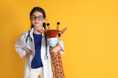 Little girl with eyeglasses, stethoscope dressed as doctor hugging toy giraffe on yellow background, space for text. Pediatrician practice
