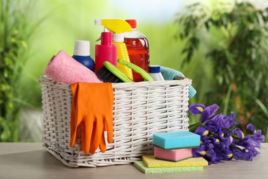 Photo of Spring cleaning. Plastic basket with detergents, supplies and beautiful flowers on wooden table outdoors