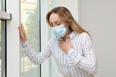 Young woman in medical mask suffering from pain during breathing near window