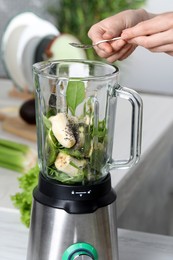 Woman adding chia seeds into blender with ingredients for green smoothie at white table, closeup