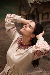 Beautiful woman wearing embroidered dress and ornate beaded necklace near old wooden well in countryside. Ukrainian national clothes