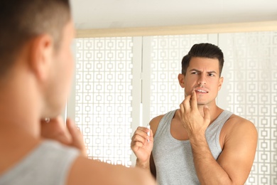 Emotional man with herpes applying cream on lips in front of mirror at home