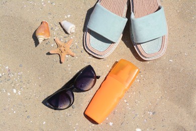 Photo of Stylish sunglasses, slippers and bottle of sunblock on sand, flat lay. Beach accessories