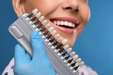 Doctor checking young woman's teeth color on blue background, closeup. Cosmetic dentistry