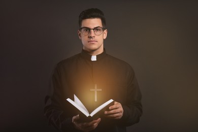 Priest in cassock holding Bible with holy light on black background