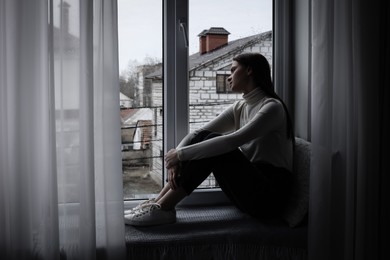 Melancholic young woman looking out of window indoors, space for text. Loneliness concept