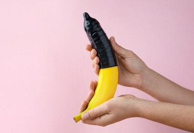 Woman holding banana in condom on pink background, closeup. Safe sex concept
