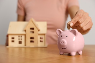 Photo of Woman putting money into piggy bank and holding house model at wooden table, focus on hand