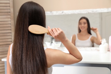 Beautiful young woman with hair brush looking into mirror in bathroom