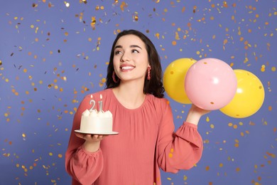 Photo of Coming of age party - 21st birthday. Smiling woman holding delicious cake with number shaped candles and balloons and looking at falling confetti on violet background