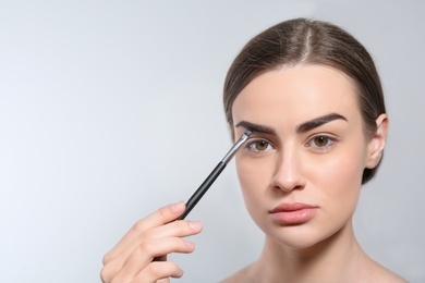 Young woman correcting shape of eyebrow with brush on light background