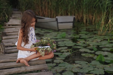 Cute little girl holding wreath made of beautiful flowers on pier near pond in evening