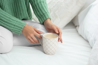 Young woman in warm sweater with hot drink on bed at home, closeup. Space for text