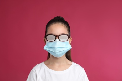 Little girl with foggy glasses caused by wearing medical face mask on pink background. Protective measure during coronavirus pandemic
