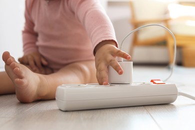 Photo of Cute baby playing with charger and power strip on floor at home, closeup. Dangerous situation