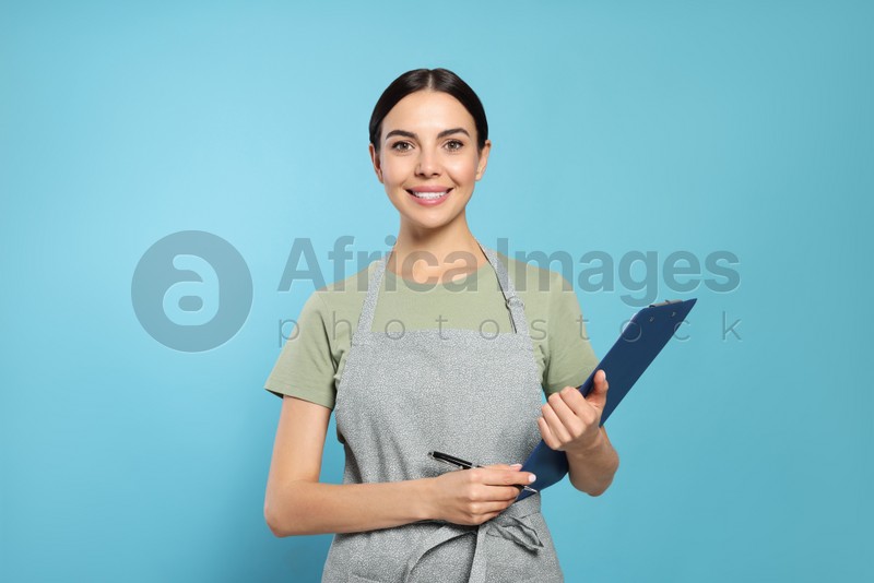 Young woman in grey apron with clipboard on light blue background