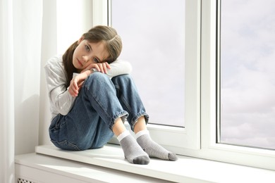 Sad little girl sitting on window sill indoors, space for text