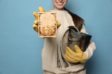 Photo of Beekeeper in uniform holding smokepot and hive frame with honeycomb on light blue background, closeup
