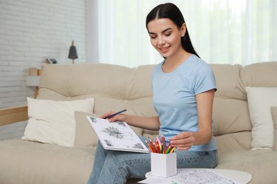 Young woman with antistress coloring page taking pencil on sofa in living room