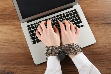 Woman with chained hands typing on laptop at wooden table, top view. Internet addiction