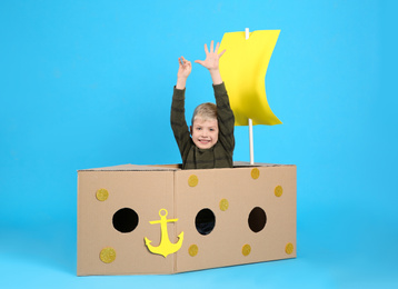 Photo of Little child playing with ship made of cardboard box on light blue background