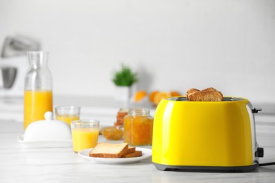 Yellow toaster with roasted bread slices, jam and glasses of juice on white marble table