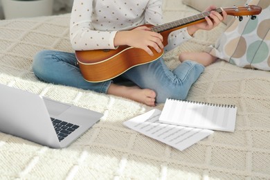 Photo of Little girl learning to play ukulele with online music course at home, closeup. Time for hobby