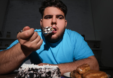 Depressed overweight man eating cake in kitchen at night