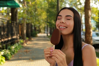 Beautiful young woman eating ice cream glazed in chocolate on city street, space for text