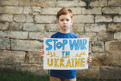 Sad boy holding poster in colors of national flag with words Stop War In Ukraine against brick wall outdoors