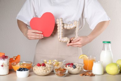 Woman holding paper heart and large intestine model near table with food, closeup. Balanced nutrition for healthy digestive system