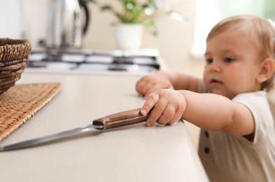 Photo of Little child touching sharp knife indoors. Dangers in kitchen