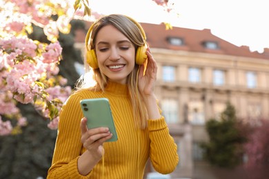 Young woman with smartphone and headphones listening to music outdoors on sunny day, space for text