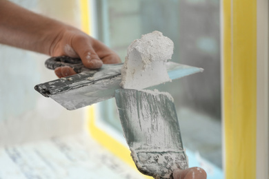 Professional worker with putty knifes and plaster near window indoors, closeup. Interior repair