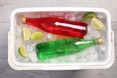 Plastic cool box filled ice cubes, lime and refreshing drinks on wooden floor, top view