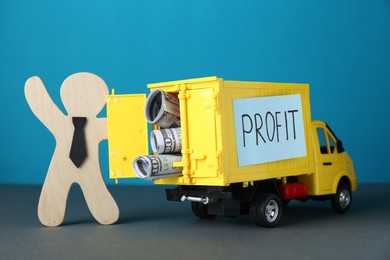 Economic profit. Wooden figure and toy truck with banknotes on grey table against light blue background